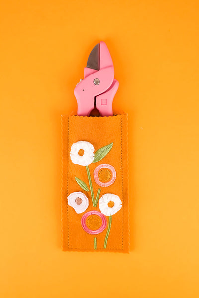 Stickmotiv Blumen für das Etui für Blumenschere in Senffarben mit einer rosafarbenen Schere halb im Etui zum Größenvergleich