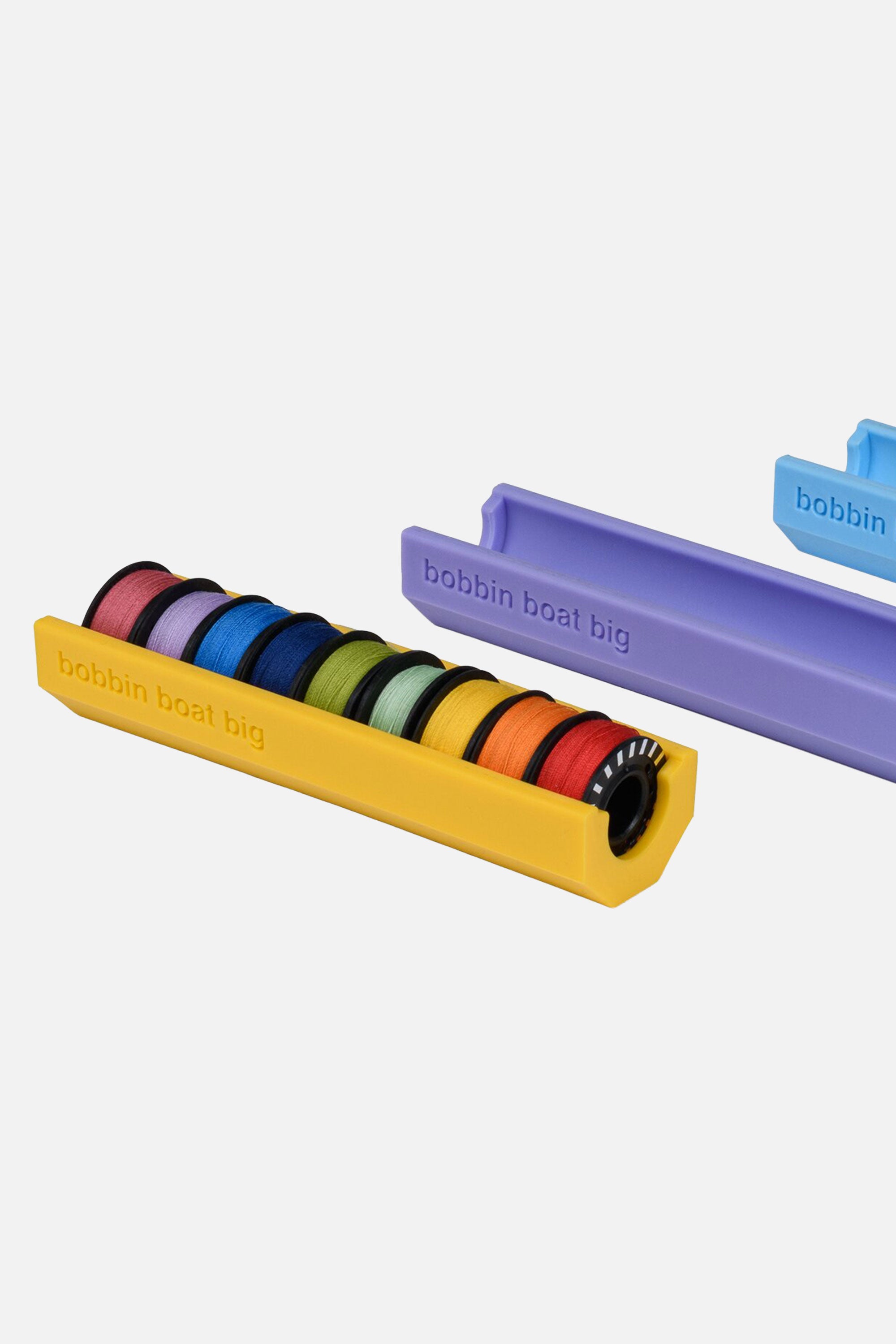 Gelber Prym Love BOBBIN BOAT BIG Spulenhalter mit eingelegten BERNINA Jumbo-Unterfadenspulen (Ø 26 mm); weitere Halter in Flieder und Blau im Hintergrund.