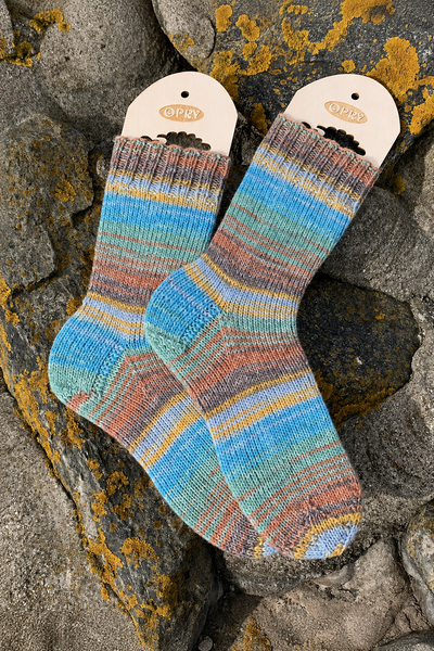 Ein Paar bunt gestrickte Socken aus 6-fädigem Garn auf einem steinigen Untergrund.
